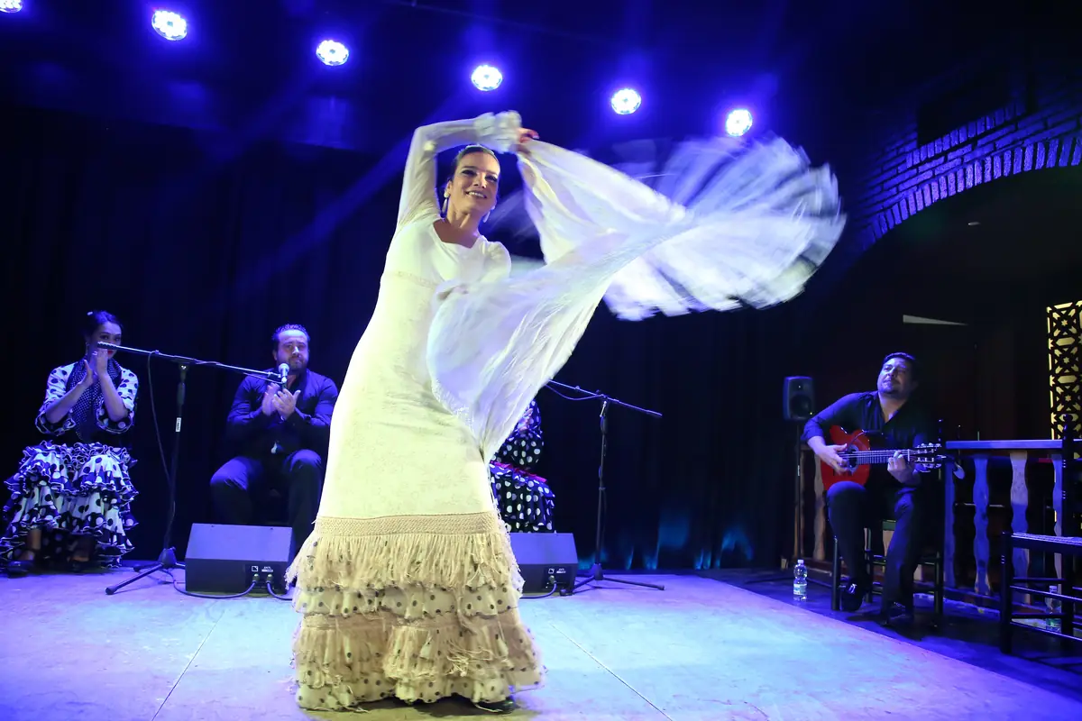 A flamenco dancer in a long white fringed dress swirls a shawl on a lit stage while a singer and a guitarist accompany her