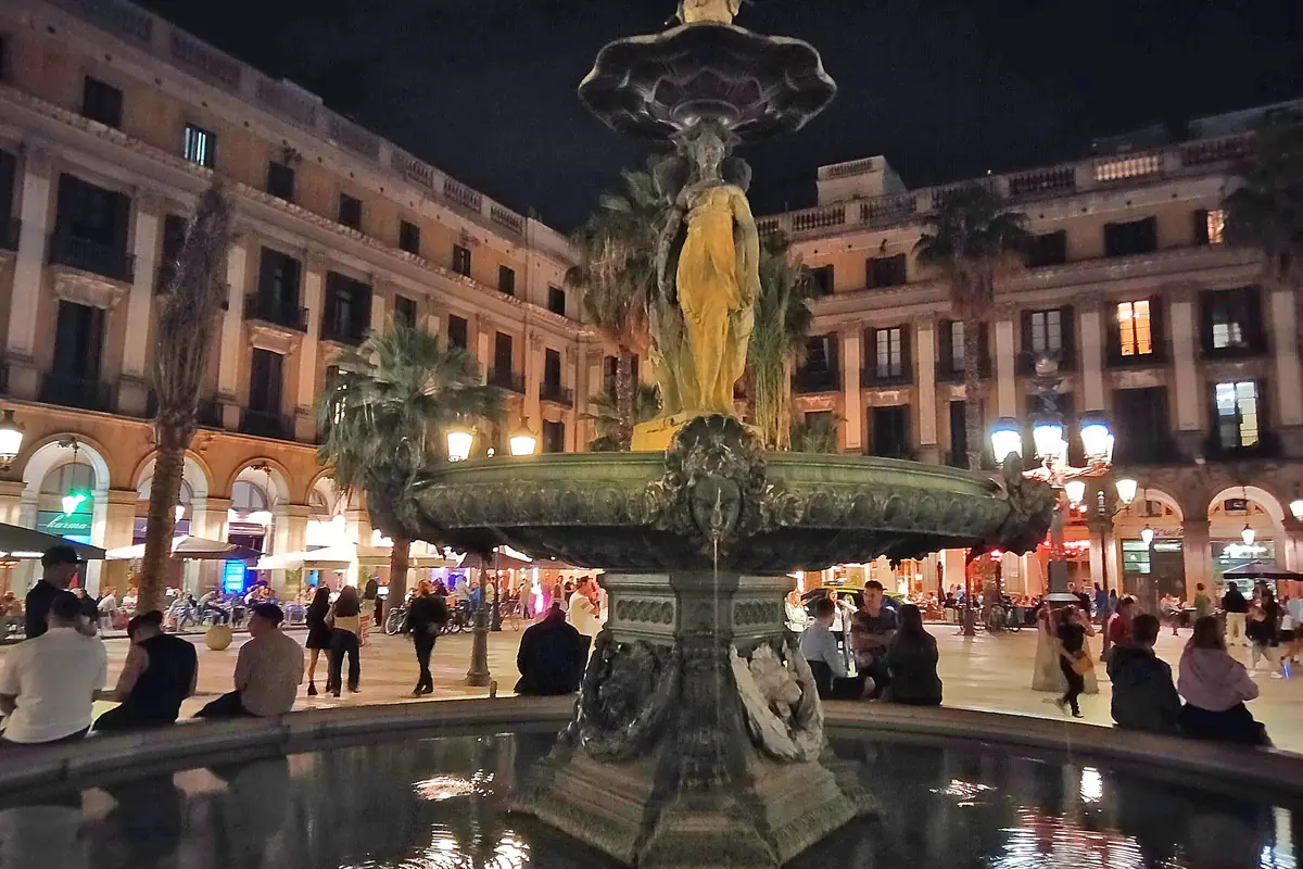The Three Graces fountain in the middle of Plaça Reial at night, lit from below, with arcaded buildings behind